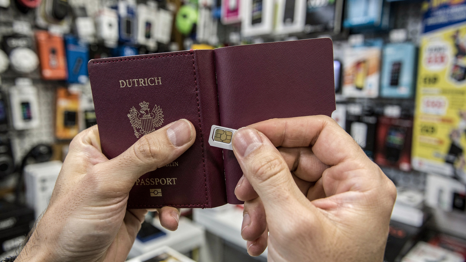 Handen van een reiziger die een paspoort en een fysieke SIM-kaart vasthouden, met op de achtergrond een balie van een telecomwinkel in een drukke Balkan stad.