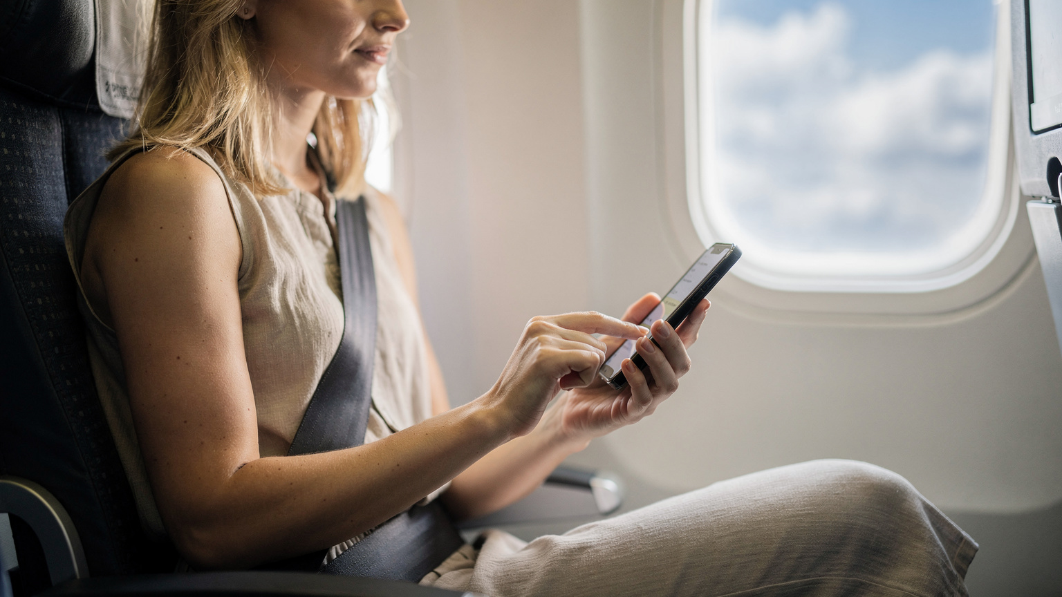 Eine junge Frau mit blonden Haaren und einem leichten Sommerkleid sitzt auf einem Fenstersitz in einem Flugzeug. Sie blickt konzentriert auf ihr modernes Smartphone und wischt mit dem Daumen über den Bildschirm, während sie die letzten Reisevorbereitungen trifft. Im Hintergrund sind durch das Flugzeugfenster sonnenbeschienene Wolken zu sehen, die auf einen bevorstehenden Urlaub hindeuten.