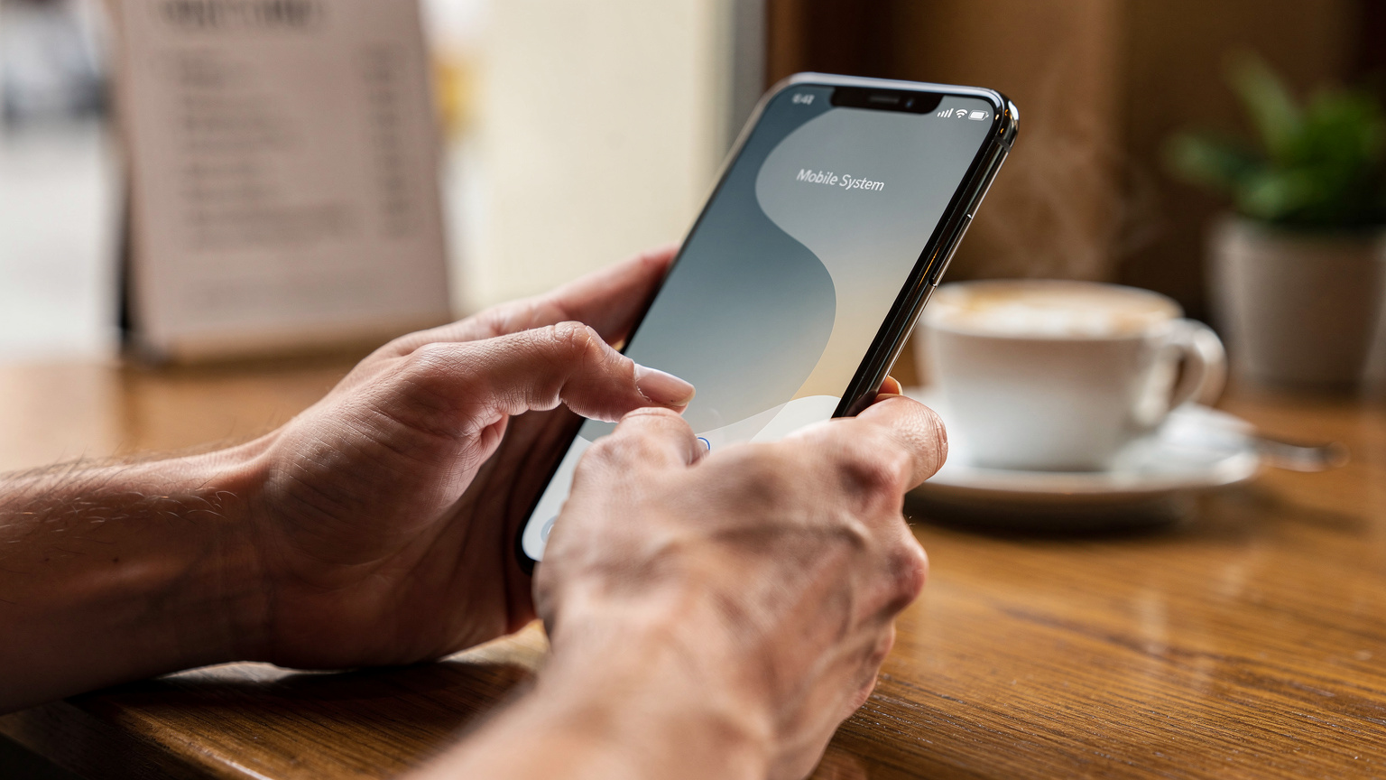 Nahaufnahme von Händen, die ein Smartphone halten und über den Bildschirm wischen, um die Mobilfunkeinstellungen zu überprüfen. Die Person sitzt an einem Holztisch in einem Café, mit einer unscharfen Tasse Kaffee im Hintergrund.