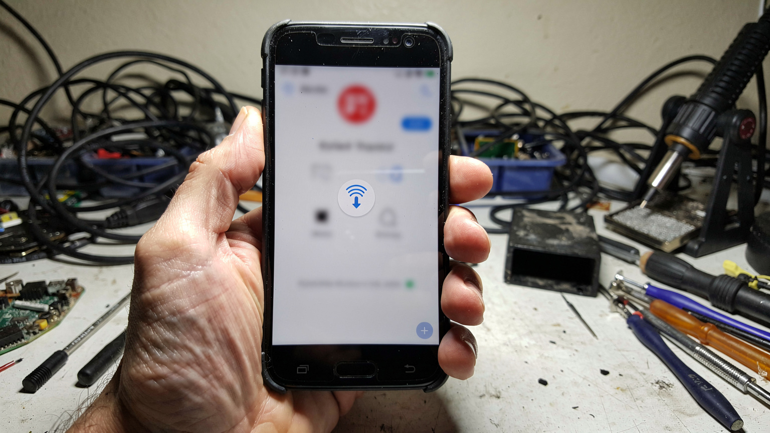 Mână de om ținând un smartphone afișând un mesaj de eroare la conectarea la rețea, cu fundalul unui birou dezordonat, plin de cabluri și unelte.