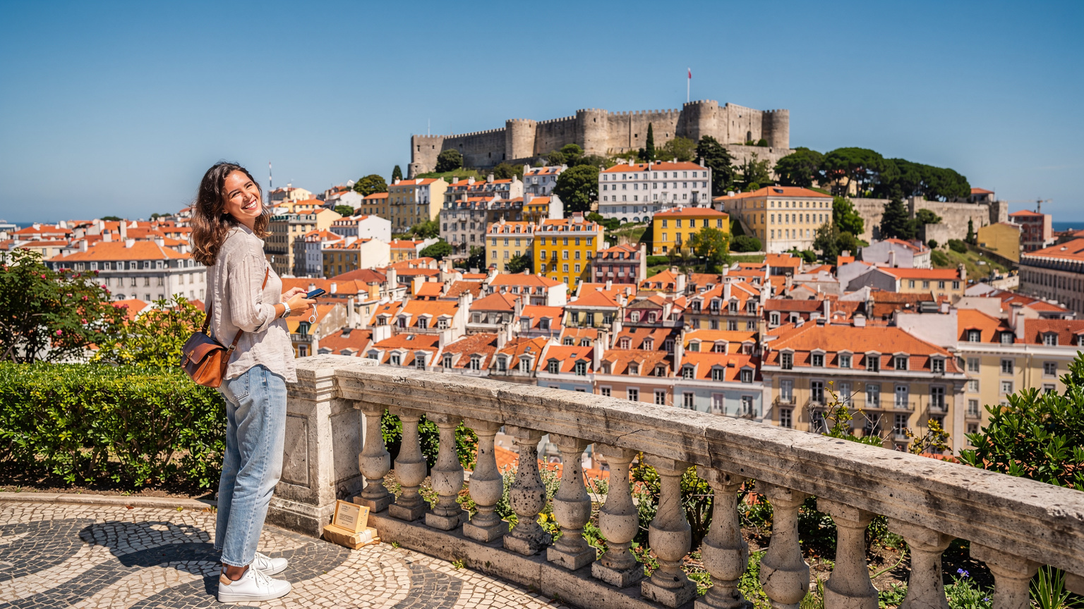 Cestovateľka s telefónom v ruke si užíva výhľad na panorámu historického centra Lisabonu z vyhliadky Miradouro de São Pedro de Alcântara.