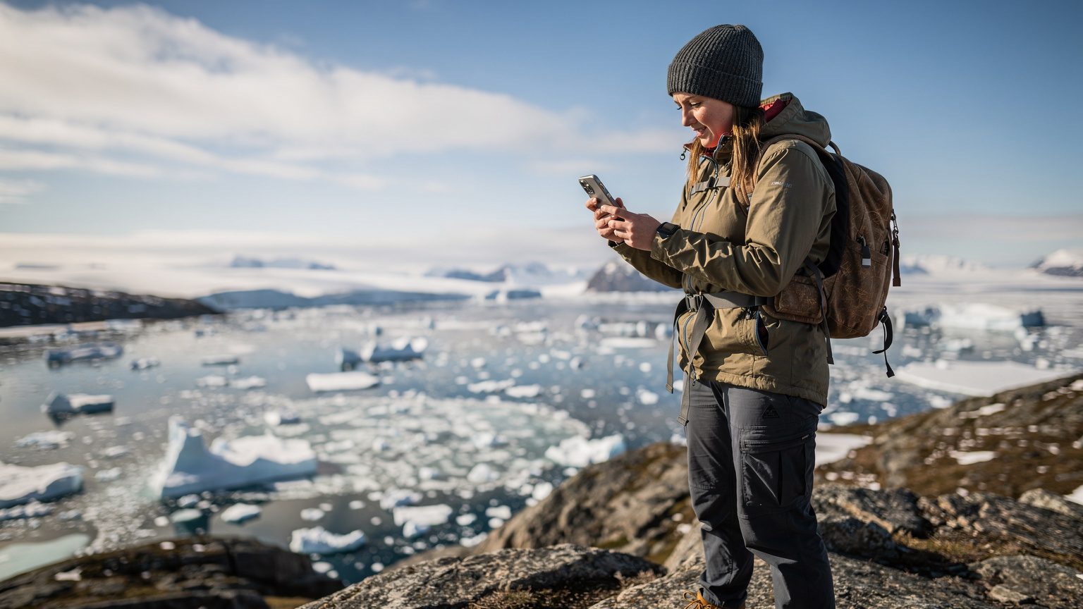 En ung rejsende tjekker sin telefon for dataforbindelse, mens hun står ved et isfjord i Grønland, hendes ansigt viser lettelse over at være online.