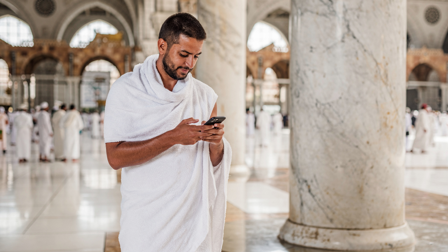 حاج يستخدم هاتفه الذكي داخل الحرم المكي الشريف، مع إضاءة طبيعية خفيفة تبرز التفاصيل المعمارية.