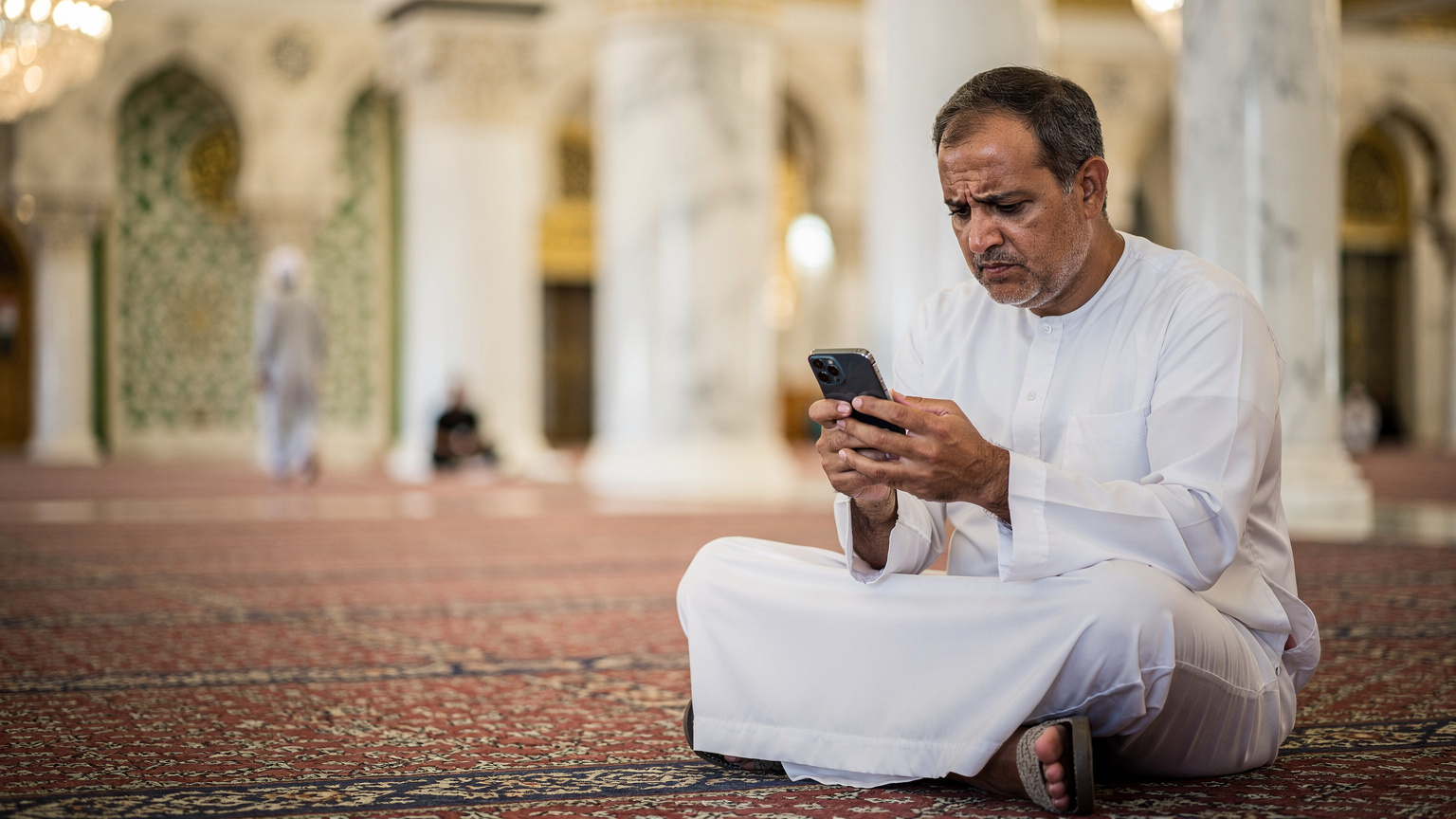 حاج جالس في منطقة هادئة داخل المسجد النبوي الشريف في المدينة المنورة، ينظر إلى هاتفه الذكي بقلق بسبب مشكلة اتصال، مع إضاءة داخلية ناعمة تبرز تعابير وجهه.