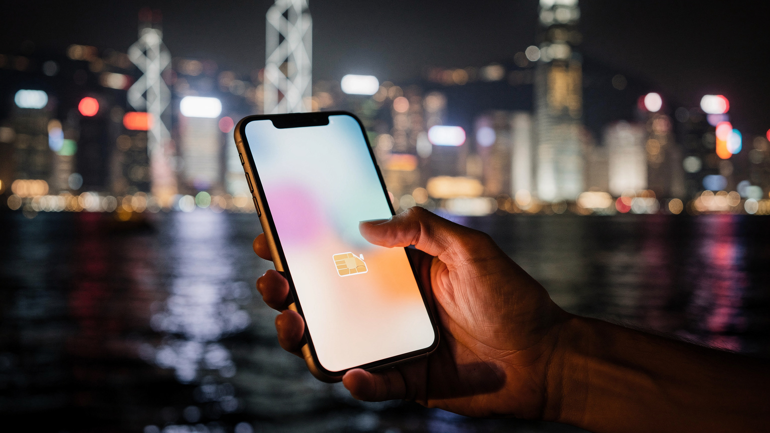 一只手正在香港维多利亚港夜景的背景下,操作 iPhone 的“蜂窝网络”设置界面,选择切换 eSIM 数据套餐,背景的灯光璀璨迷人。