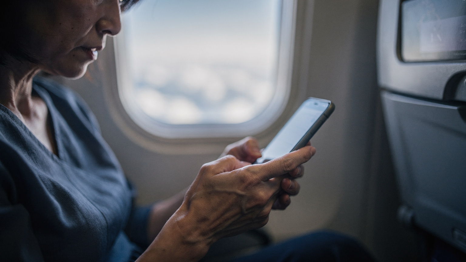 スマートフォンを手に持ち、困った表情で画面を見つめる旅行者。隣には飛行機の窓から見える雲が映り、機内での通信トラブルを暗示している。
