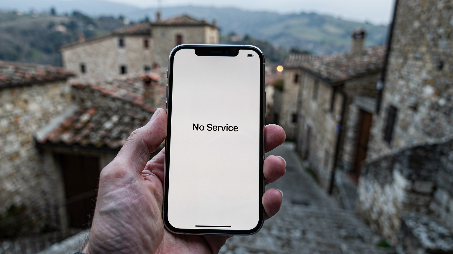 Ένα smartphone εμφανίζει ένα μήνυμα 'No Service' ενώ κρατιέται από ένα χέρι σε ένα γραφικό, απομακρυσμένο ορεινό χωριό της Τοσκάνης, με παλιά πέτρινα κτίρια και στενά σοκάκια στο βάθος.