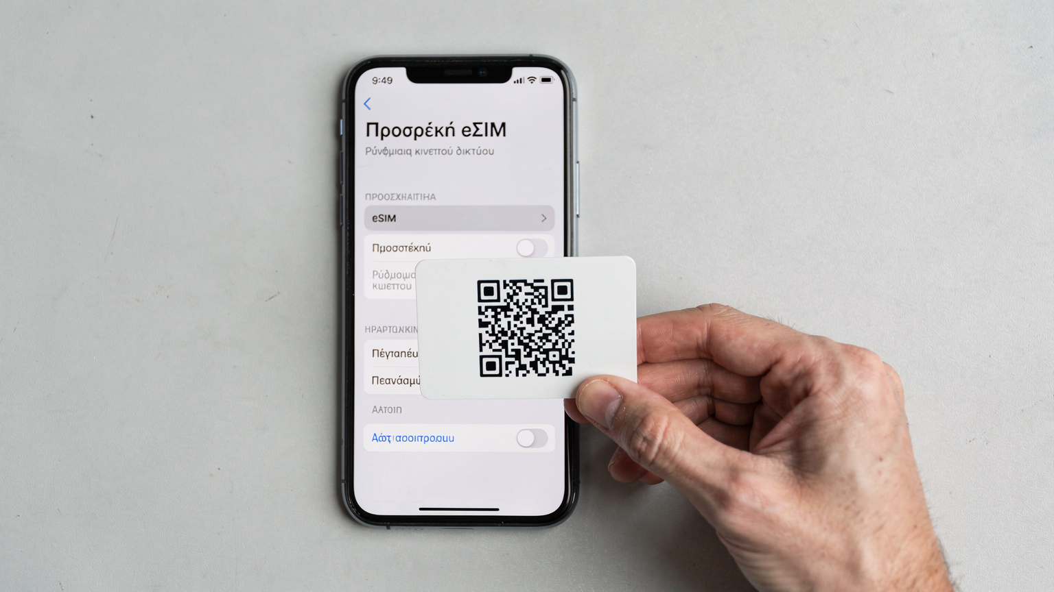 Ένα smartphone iPhone εμφανίζει τις ρυθμίσεις για την προσθήκη eSIM, με το χέρι ενός ατόμου να κρατάει μια κάρτα με έναν κωδικό QR έτοιμο για σάρωση.