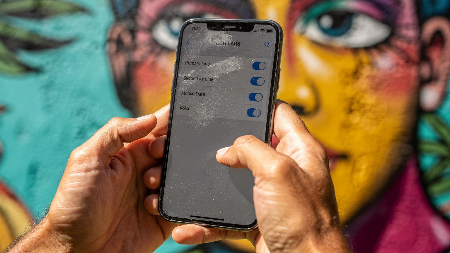 Close-up of hands holding a smartphone, the screen showing phone settings with 'Primary' and 'Secondary' lines, set against a vibrant background of a street art mural in Rio de Janeiro.