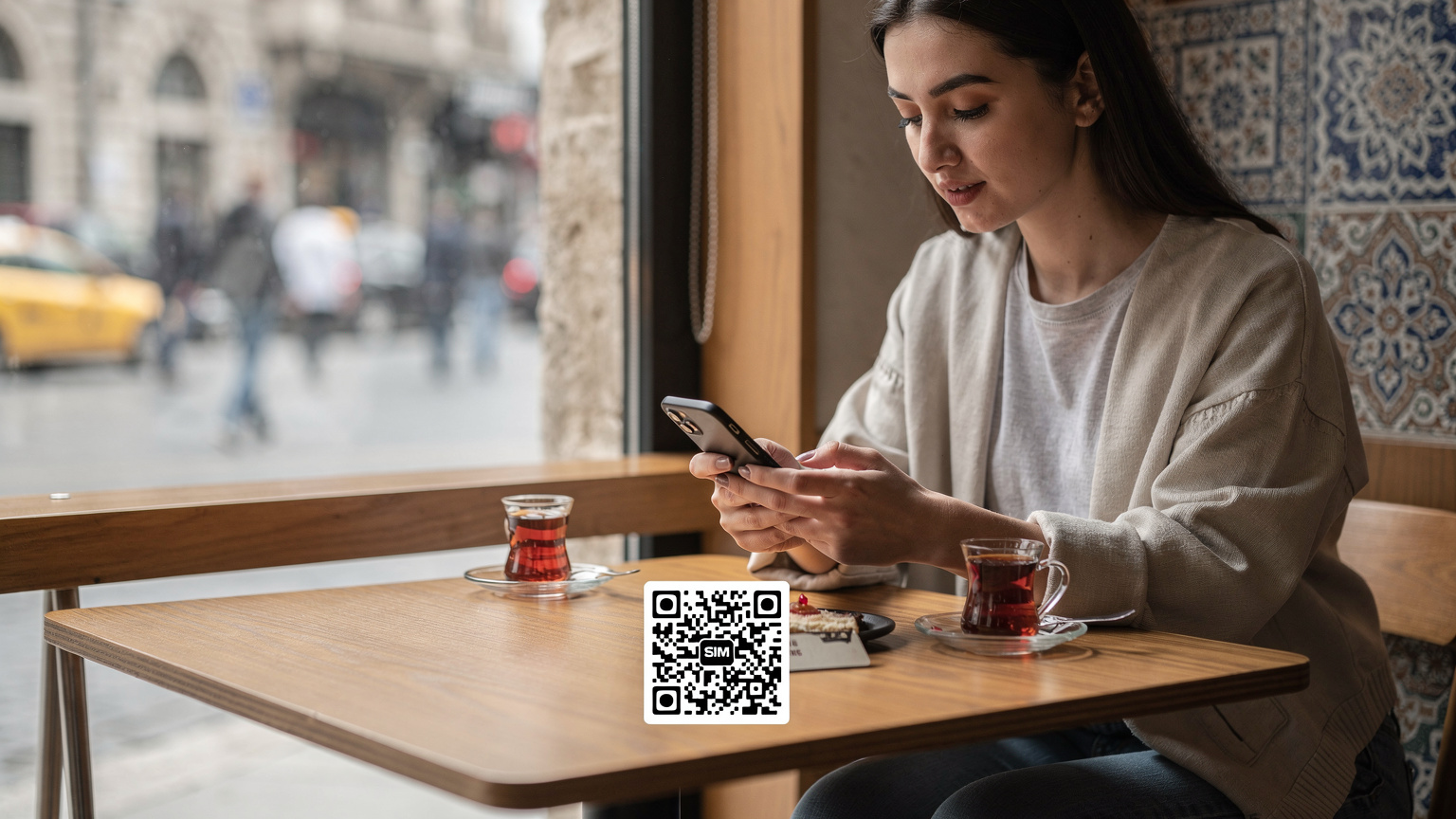 İstanbul'daki modern bir kafede, pencereden gelen yumuşak doğal ışık altında, genç bir gezgin elindeki akıllı telefonunu kullanarak Cellesim eSIM'inin QR kodunu tararken gösteriliyor.