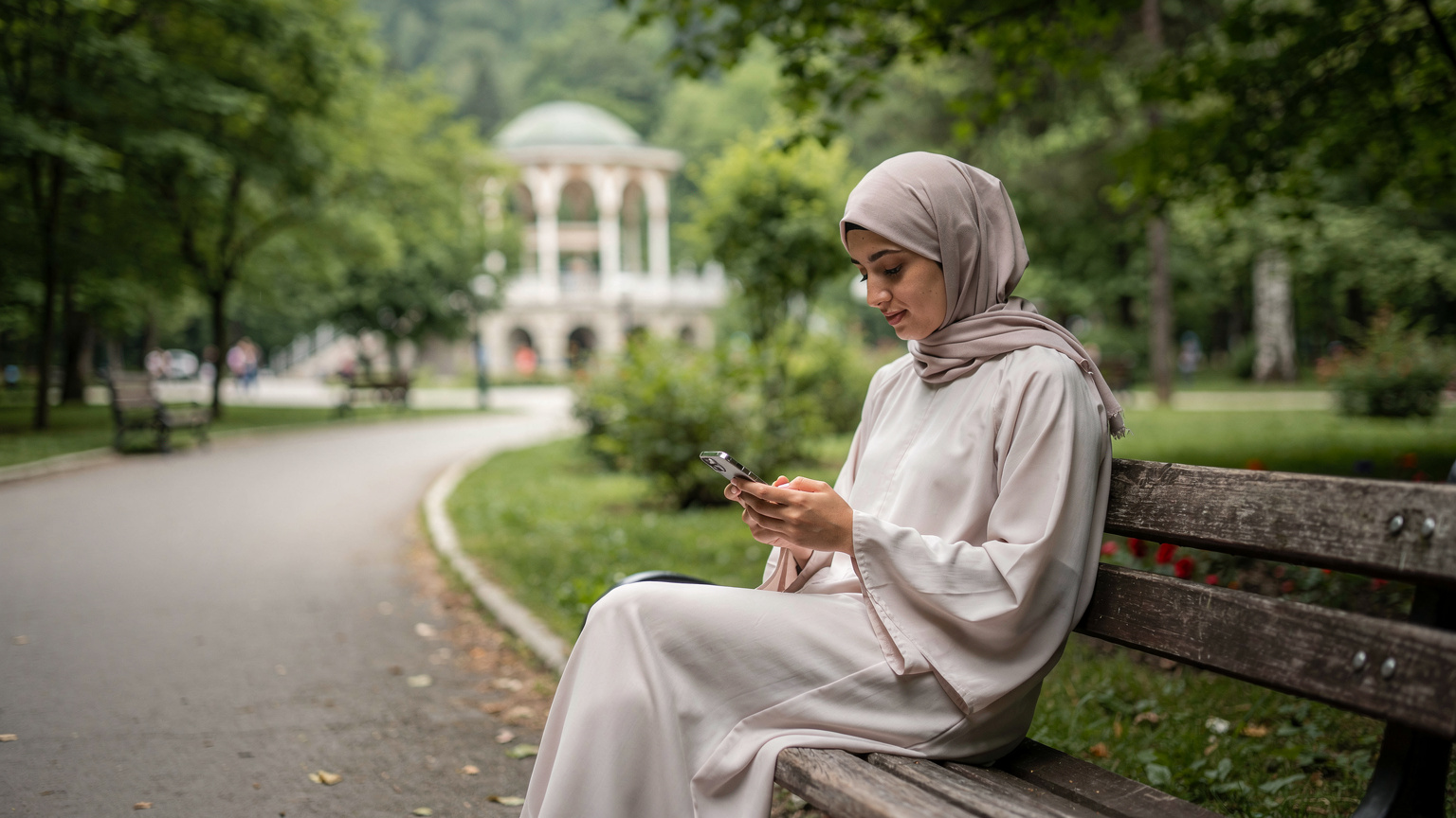 امرأة خليجية شابة تجلس على مقعد خشبي في حديقة بورجومي بجورجيا, وتستخدم هاتفها بتركيز شديد.