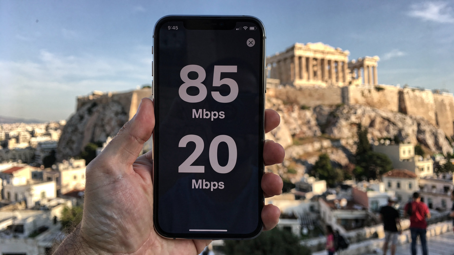 A close-up of a smartphone screen displaying a speed test result, showing high download and upload speeds, held against a backdrop of the Acropolis.