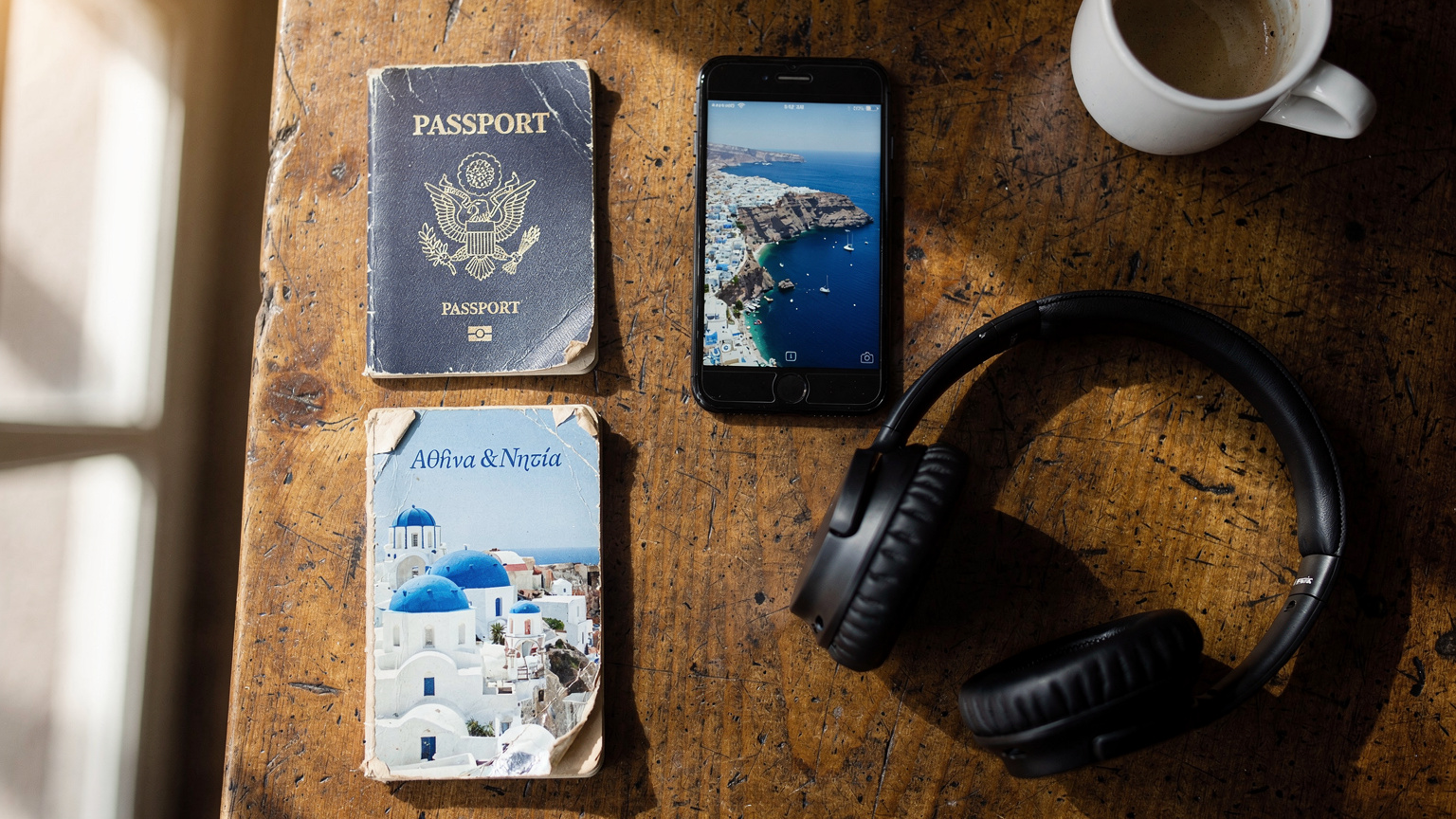 An overhead flat-lay of travel essentials: a passport, a smartphone displaying a Greek map, noise-canceling headphones, and a small travel guide, all neatly arranged on a rustic wooden table in a café.