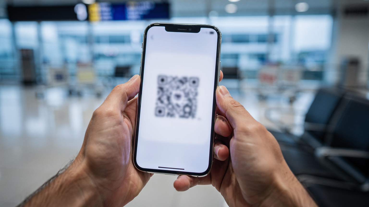 Крупний план рук, які тримають смартфон, на екрані якого відображається QR-код для активації eSIM. На задньому плані, розмито, видно елементи аеропорту, такі як багажні візки та вивіски, створюючи відчуття транзиту та швидкої потреби у зв'язку.