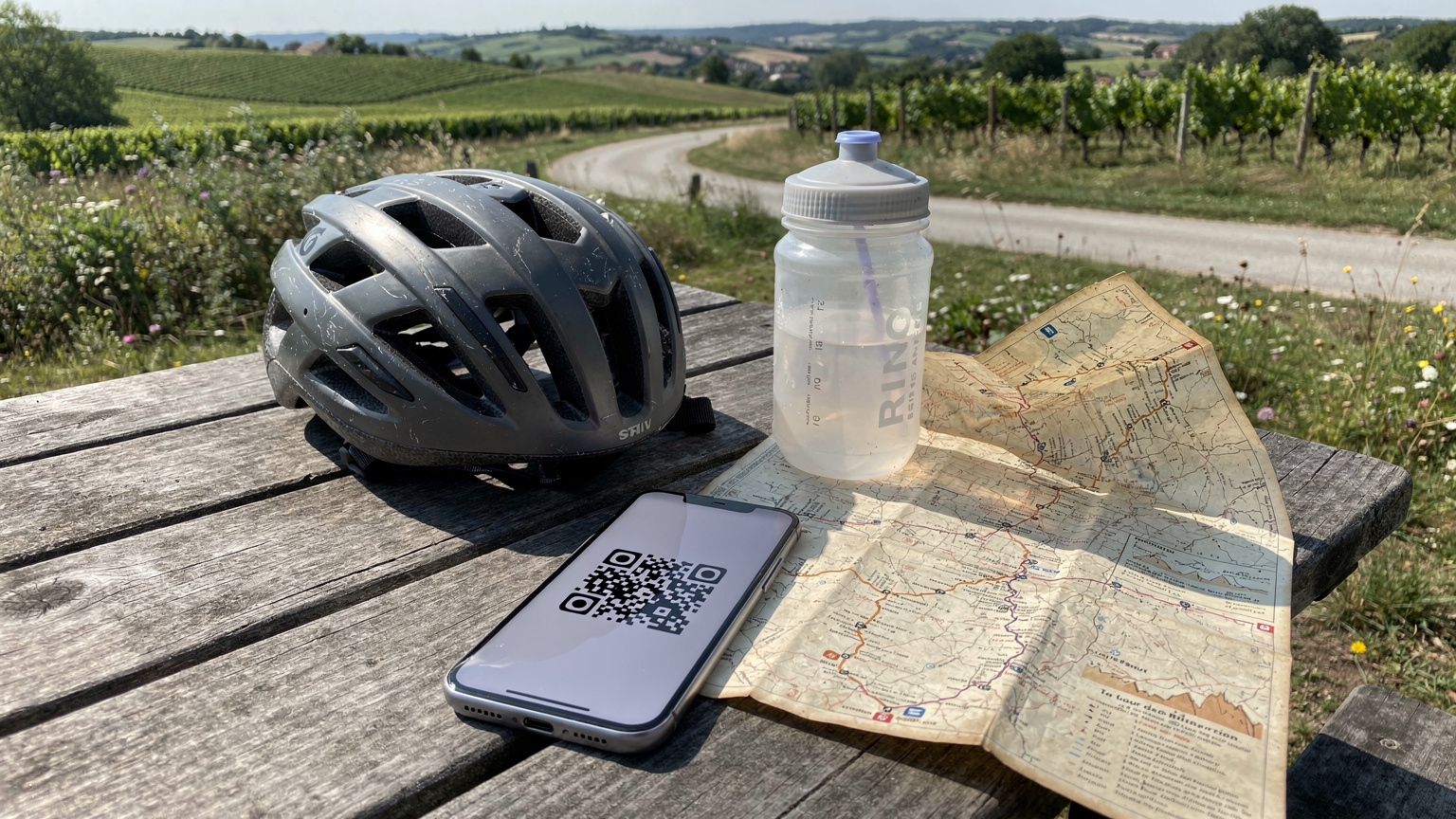 Een gedetailleerde weergave van een smartphone die een QR-code toont voor eSIM-installatie, liggend op een picknicktafel naast een fietshelm, bidon en een gedrukte kaart van de Tour de France route, met een landelijk Frans landschap op de achtergrond.