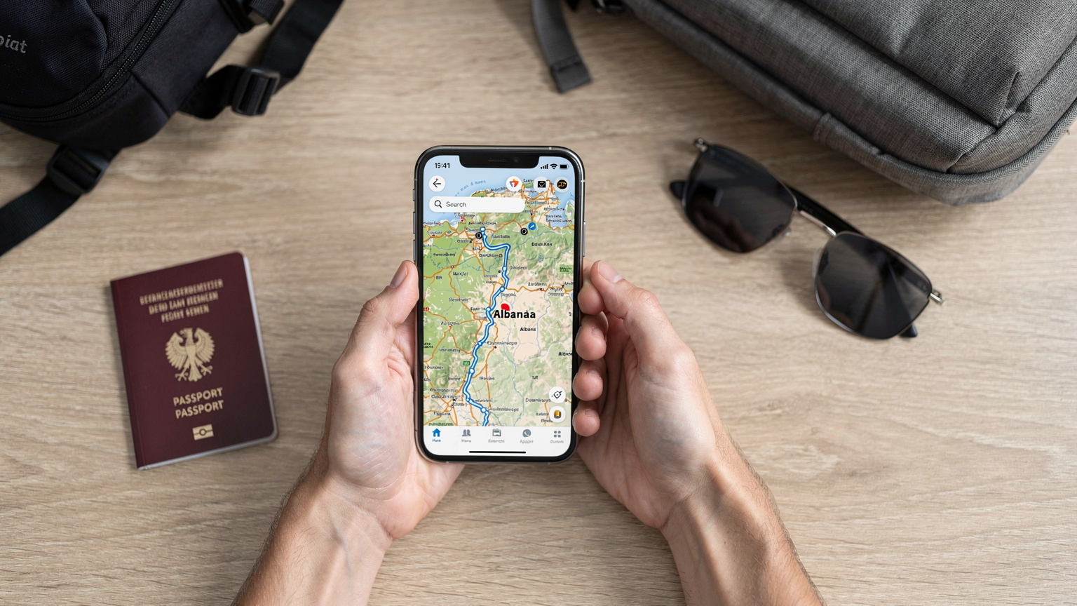 Hände halten ein Smartphone, das eine Reise-App mit einer Albanien-Route anzeigt, neben einem Reisepass, einer Sonnenbrille und einem kleinen Rucksack auf einem Holztisch, bereit zur Abfahrt.
