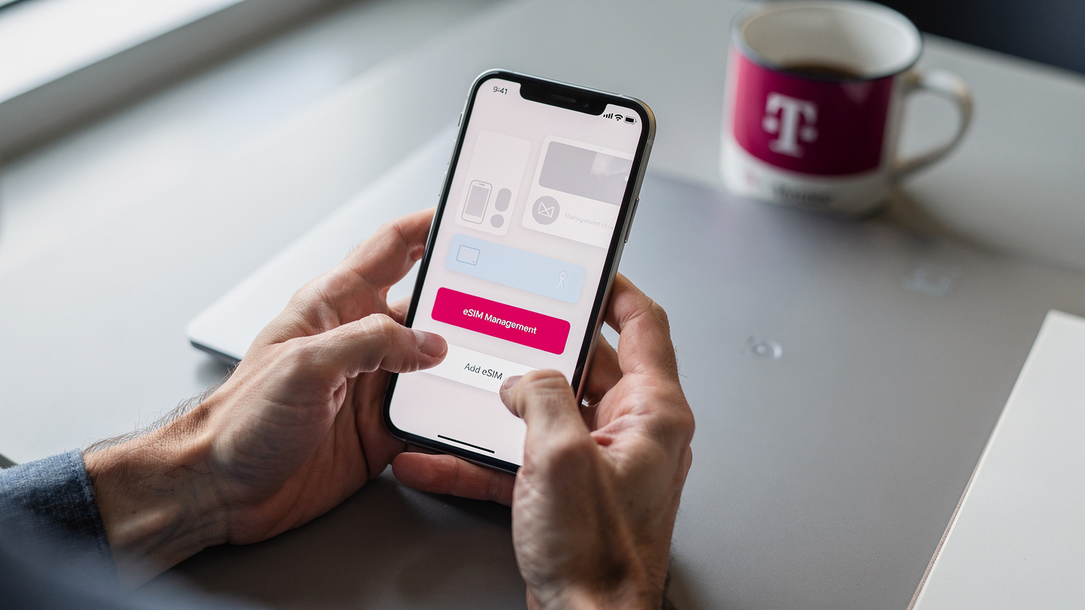 A close-up of a smartphone screen displaying a T-Mobile app with an option for eSIM management, with blurred hands interacting with the device.
