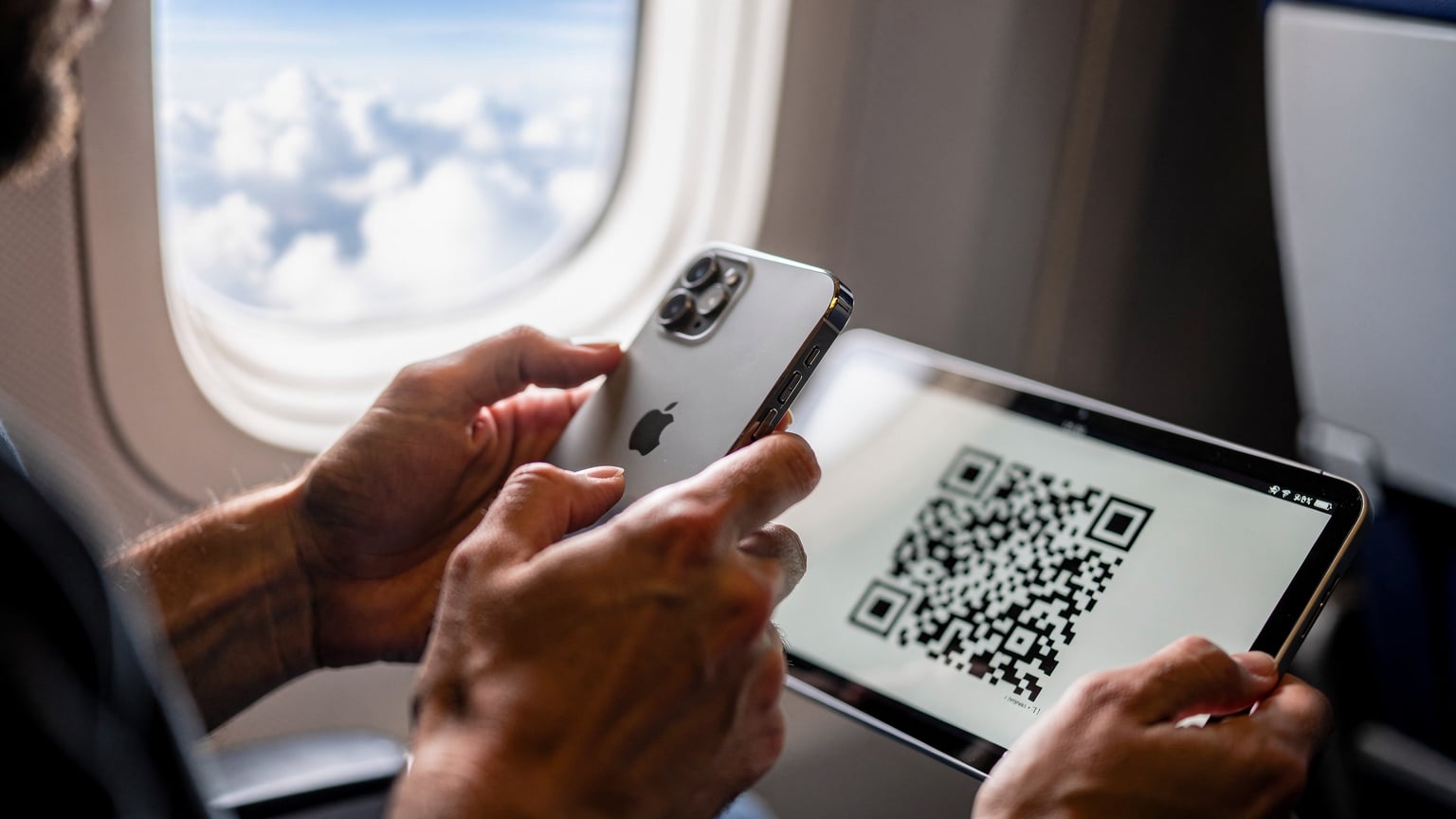 Tangan seorang traveler memegang iPhone 17, sedang memindai kode QR eSIM Cellesim yang ditampilkan di layar tablet lain, dengan latar belakang jendela pesawat yang menunjukkan awan.