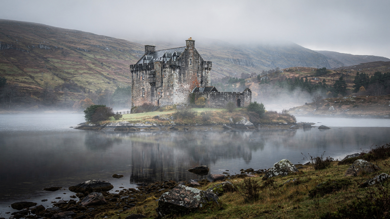 Un majestuoso castillo escocés, cubierto de niebla, se alza sobre un lago, evocando la rica historia del Reino Unido.