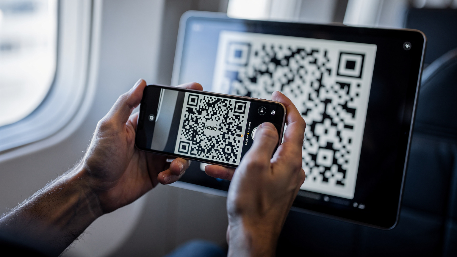 Zbliżenie na dłonie rodzica trzymającego iPhone'a 18 i skanującego kod QR eSIM z ekranu tabletu, w tle rozmazane wnętrze samolotu.