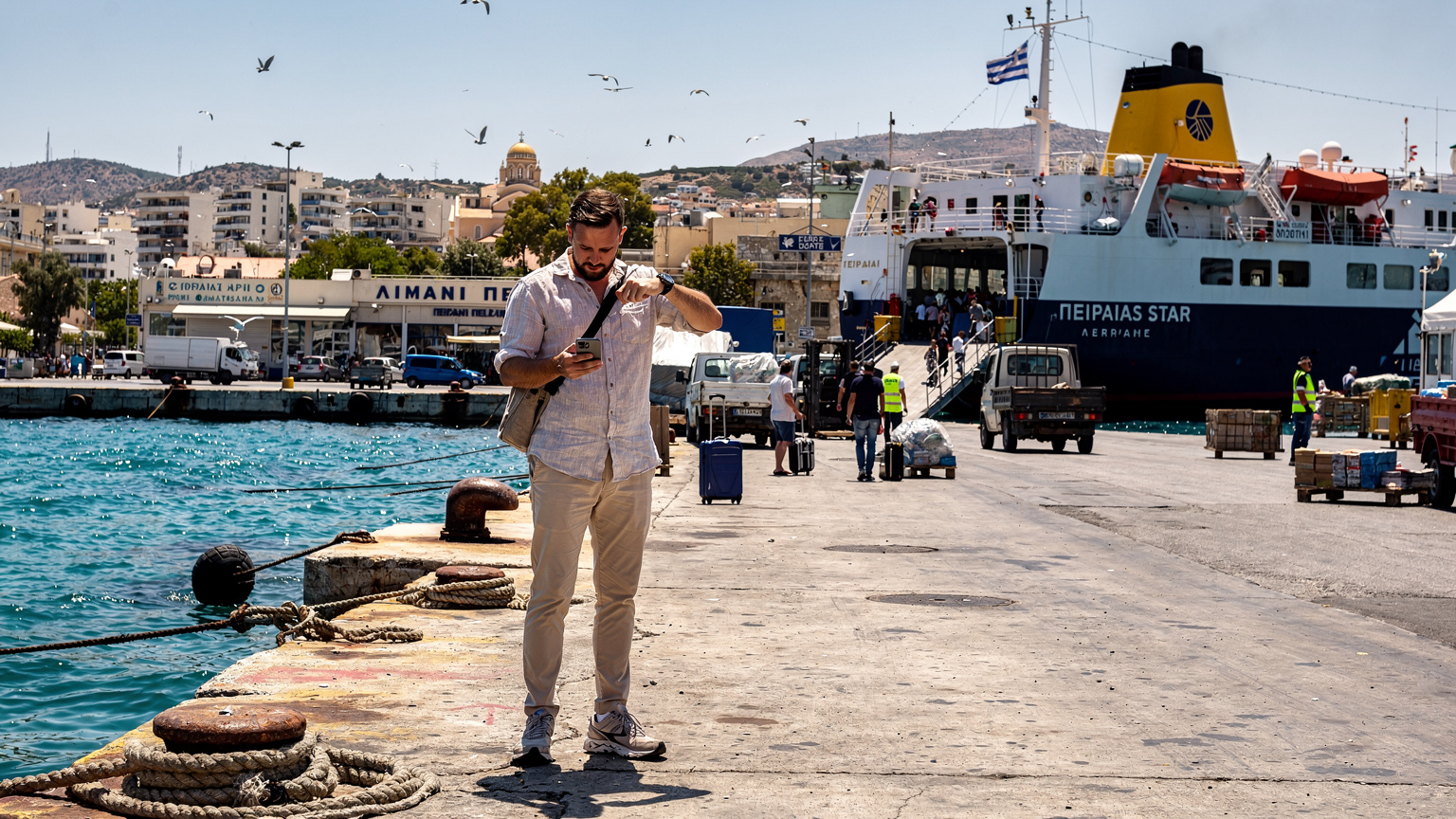 En rejsende tjekker sin telefon for færgeafgange fra havnen i Piraeus, med en traditionel græsk færge i baggrunden.