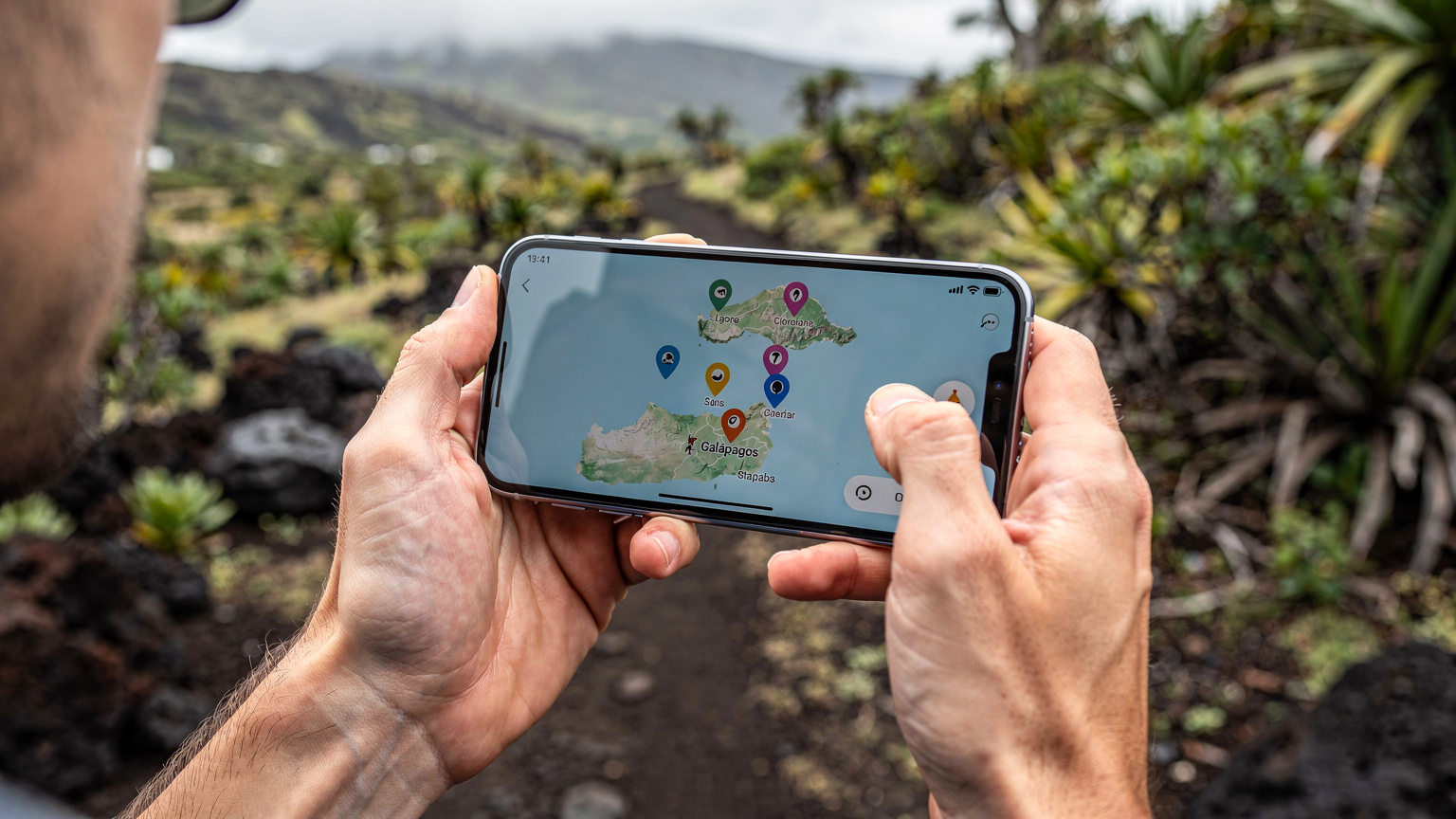 Des mains tiennent un smartphone affichant une application de cartographie hors ligne, avec des épingles marquant des sites d'intérêt aux Galápagos. L'arrière-plan flou suggère un sentier de randonnée avec de la végétation tropicale.