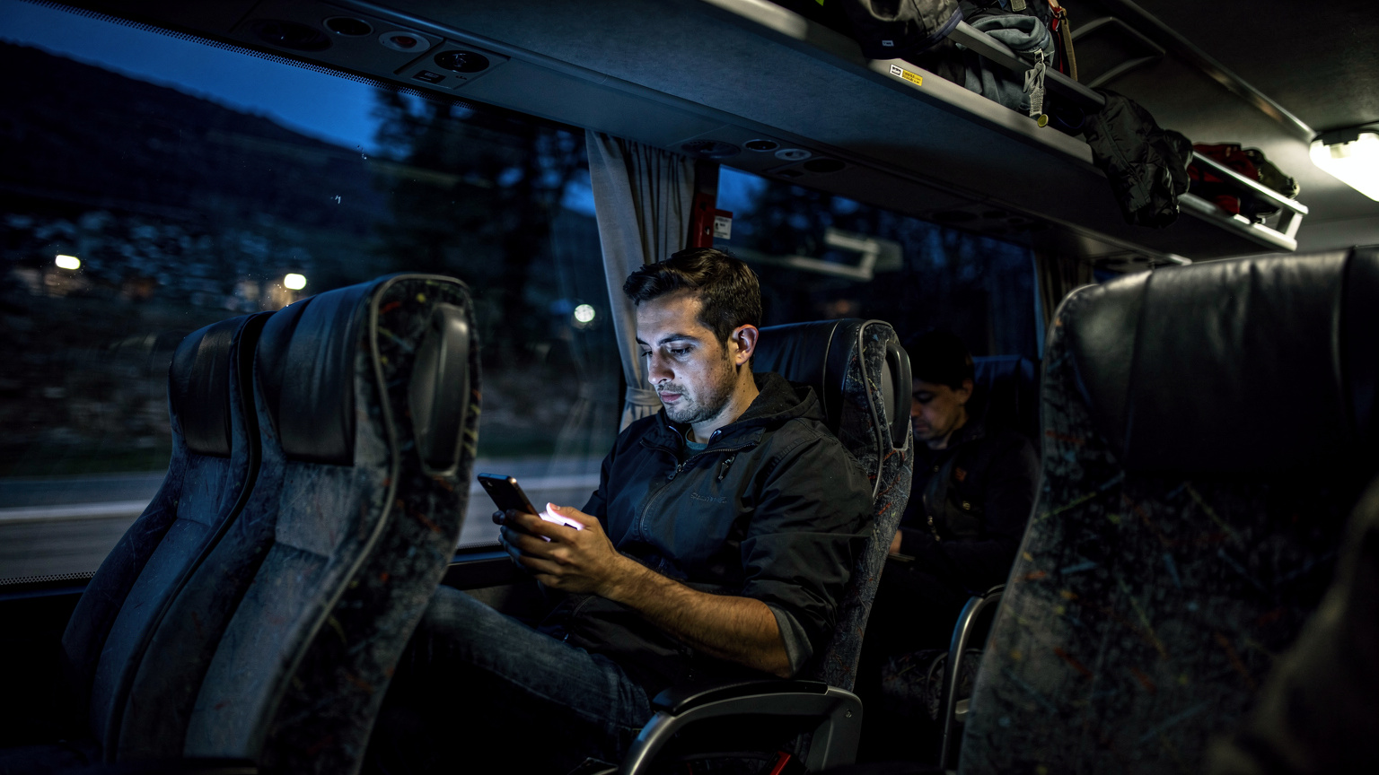 Ein Traveller in einem Nachtbus auf dem Balkan, der besorgt auf sein Smartphone schaut, das keinen Empfang anzeigt, während draußen die dunkle Landschaft vorbeizieht.