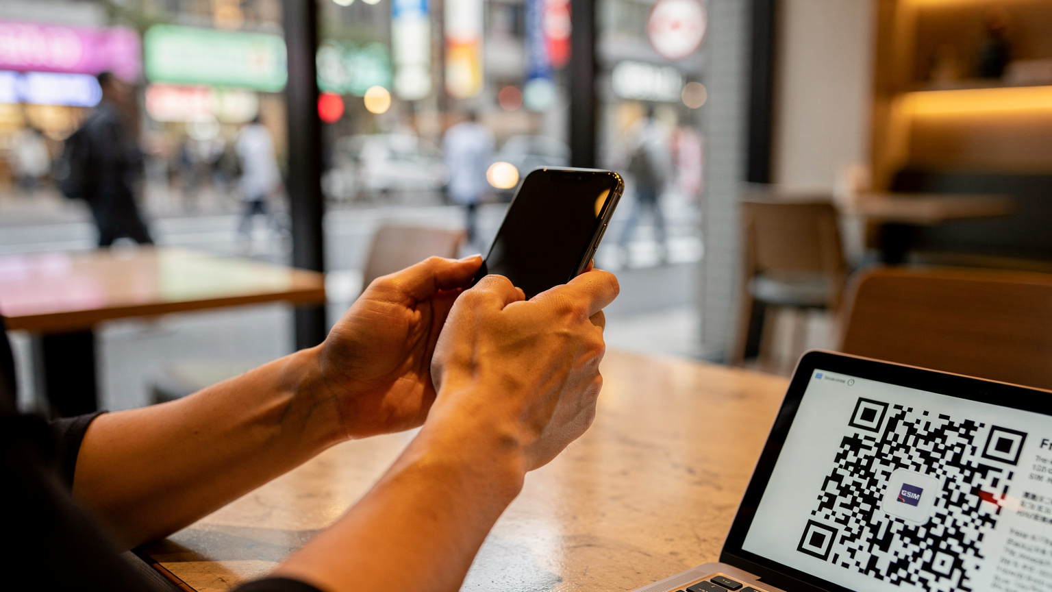 Des mains tenant un iPhone, scannant un QR code d'eSIM dans un café animé du quartier de Shinjuku à Tokyo, avec des passants flous en arrière-plan.