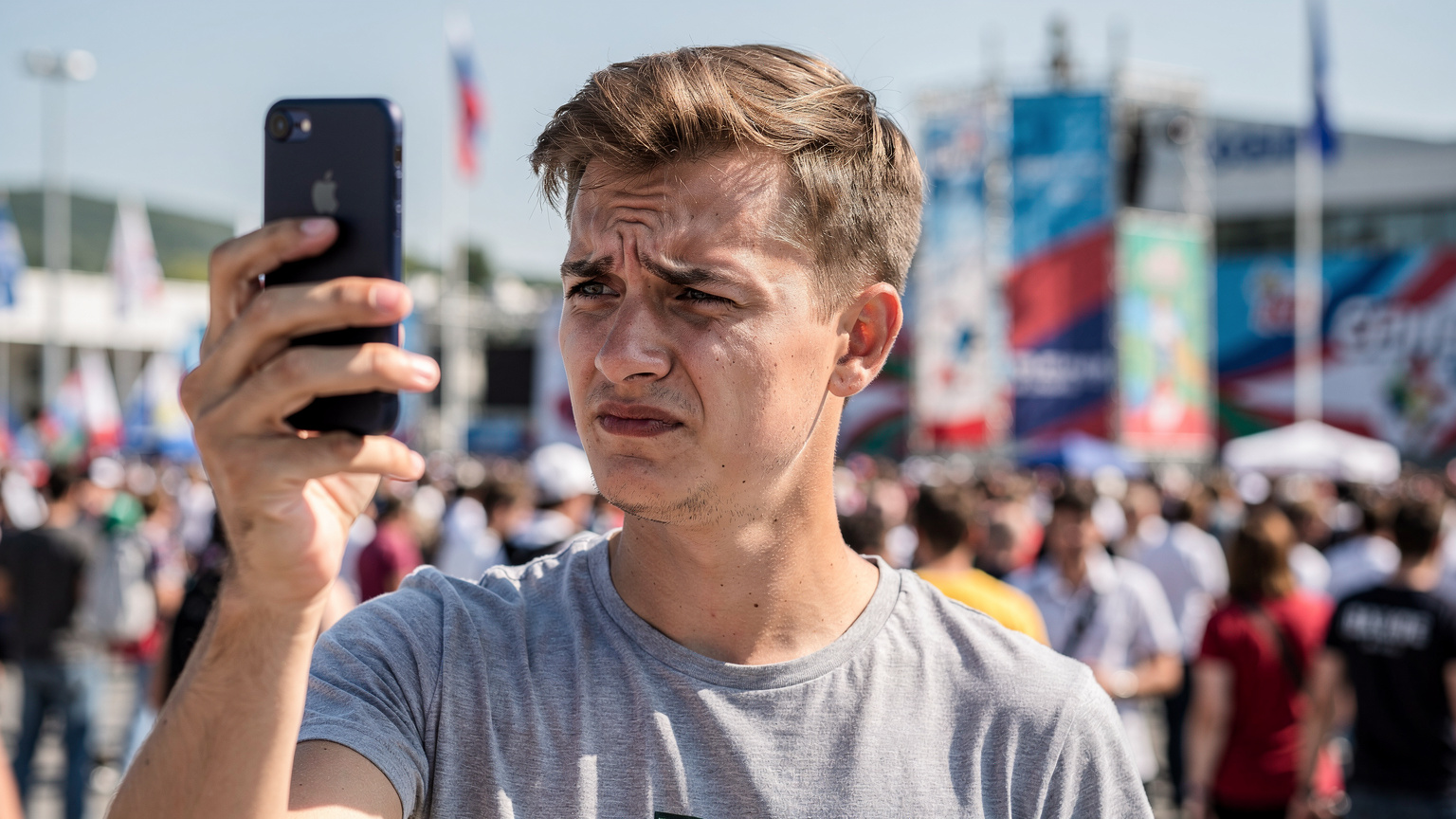 Молодой человек с озабоченным выражением лица держит смартфон, пытаясь подключиться к сети на переполненной площади, символизируя проблемы со связью на массовых мероприятиях.