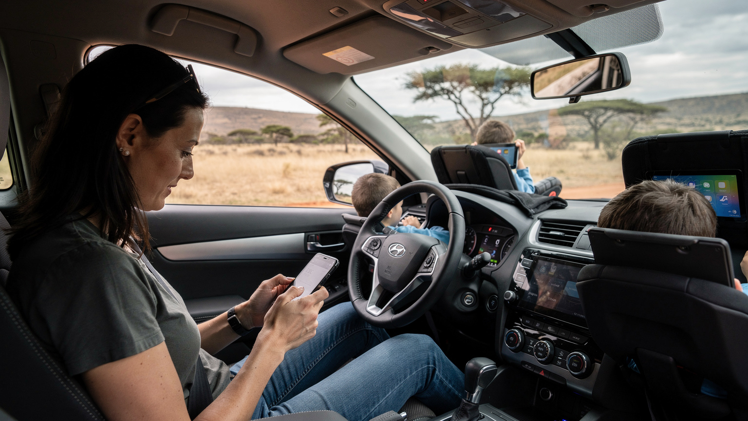Een moeder installeert rustig een eSIM op haar smartphone terwijl haar twee kinderen geconcentreerd bezig zijn met hun tablets op de achterbank van een auto, geparkeerd bij een typisch Zuid-Afrikaans landschap.
