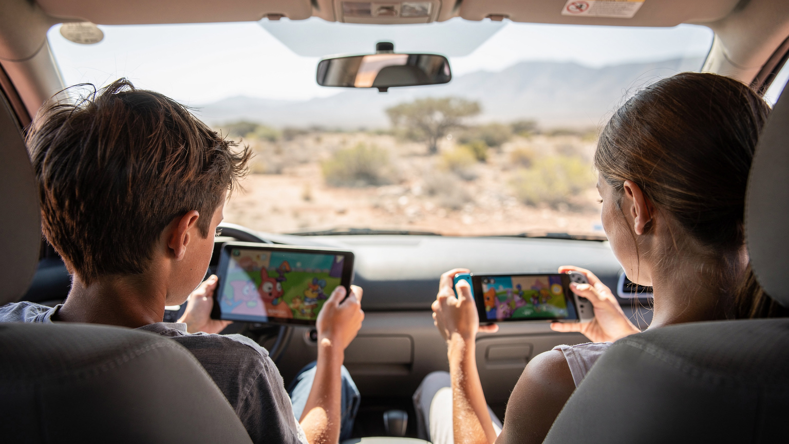 Twee kinderen zitten geconcentreerd op de achterbank van een auto, de één met een tablet waarop een tekenfilm speelt, de ander met een draagbare spelcomputer, terwijl ze door een typisch Zuid-Afrikaans landschap rijden.
