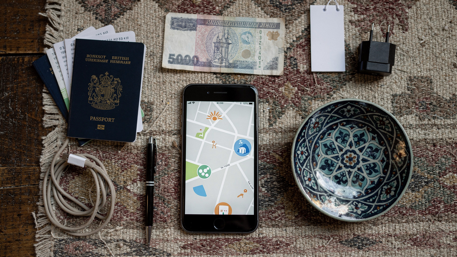 An overhead flat-lay of travel essentials for Central Asia: a British passport, a smartphone displaying a map application, a Uzbekistani Som banknote, and a small, traditional ceramic souvenir.