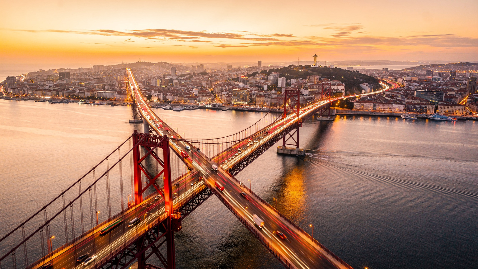 Uma vista aérea da Ponte 25 de Abril em Lisboa ao pôr do sol, com os carros passando e o Rio Tejo em tons quentes. Uma cena que evoca a beleza e a conectividade de Portugal.