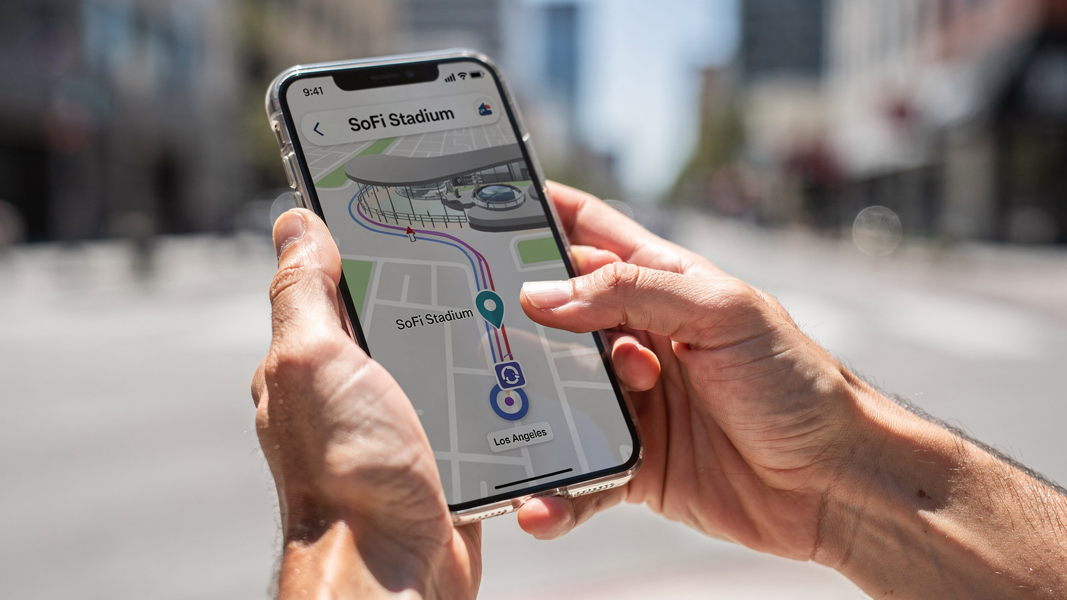 Mãos de uma pessoa segurando um smartphone, mostrando um aplicativo de mapa com uma rota destacada para o SoFi Stadium em Los Angeles, com o dedo indicador deslizando na tela.