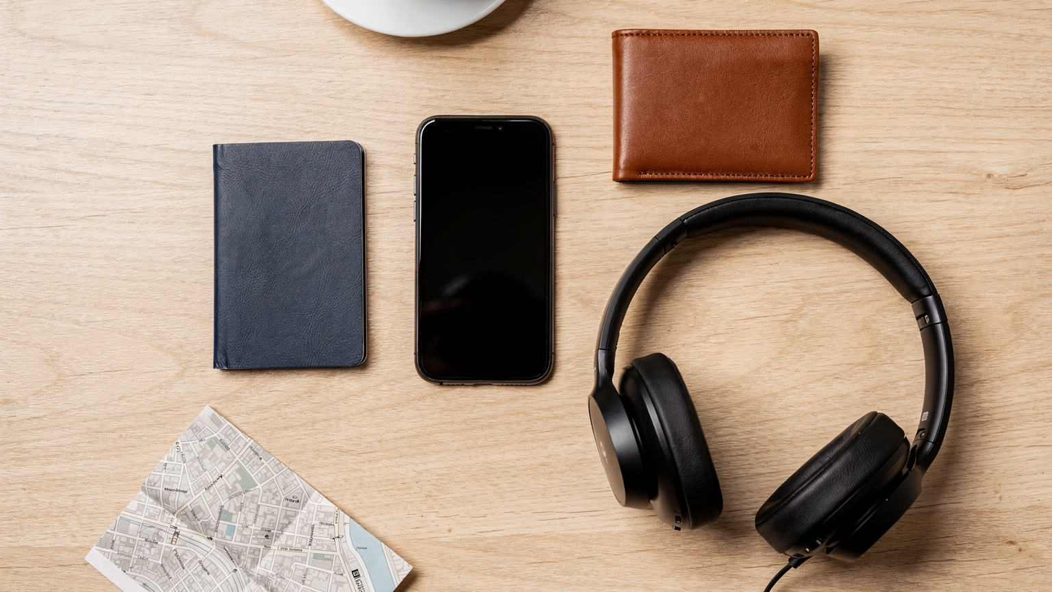 An overhead flat-lay shot of essential travel items: passport, smartphone, wallet, noise-cancelling headphones, and a small, folded map, all neatly arranged on a cafe table.