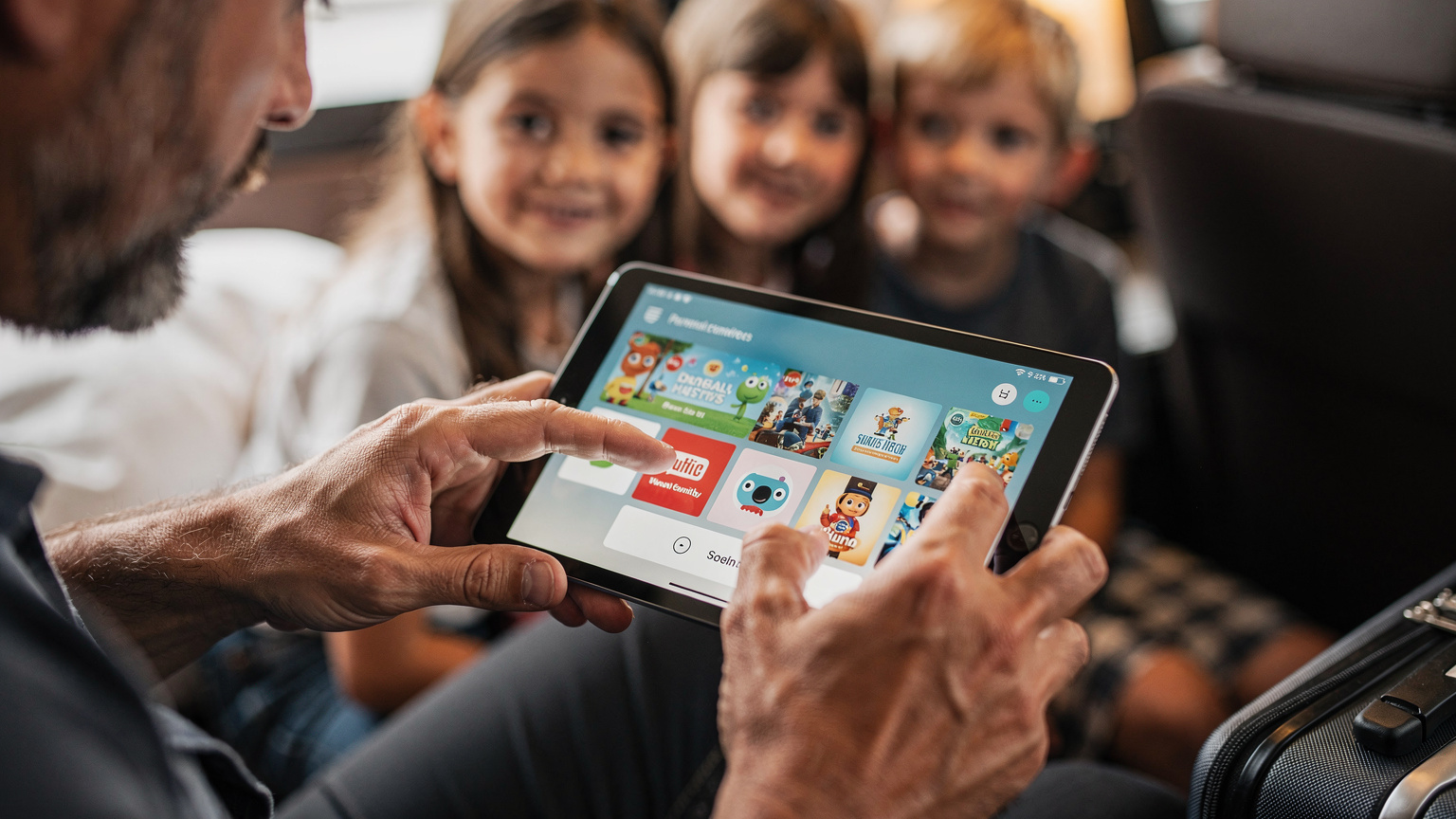 Nahaufnahme der Hände eines Vaters, der auf einem Tablet für seine zwei Kinder eine kindersichere Oberfläche einrichtet oder eine Serie zum Offline-Schauen herunterlädt. Man erkennt im Hintergrund verschwommen die Gesichter der Kinder, die gespannt zuschauen. Der Vater ist konzentriert auf das Tablet, das eine bekannte Streaming-App zeigt.