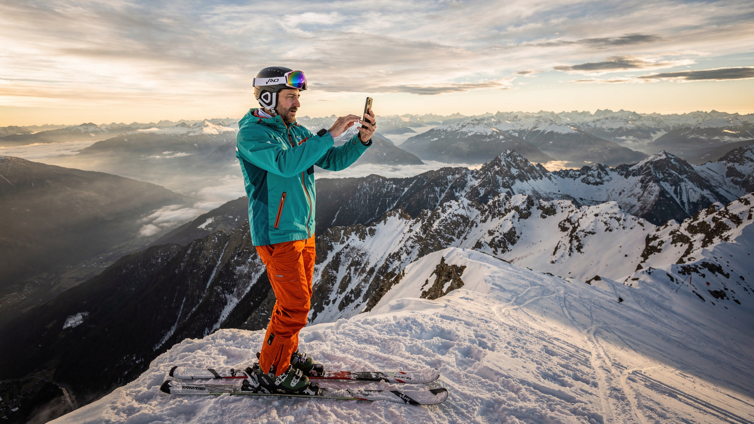 Ein Skifahrer in voller Ausrüstung steht auf einem schneebedeckten Berggipfel in den Pyrenäen von Andorra und versucht, mit seinem Smartphone ein Foto der majestätischen Landschaft zu machen, während er mit einer Hand eine Nachricht tippt. Die Abendsonne taucht die Szene in ein warmes Licht und betont die Weite der Berge.