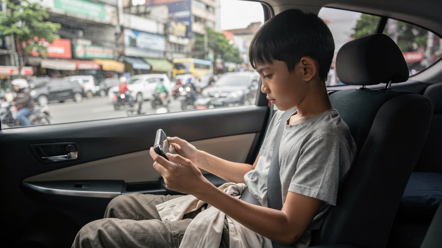 Seorang remaja sekitar 12 tahun sedang asyik bermain game di konsol Nintendo Switch miliknya di kursi belakang mobil, dengan pemandangan jalanan kota yang sibuk di luar jendela.