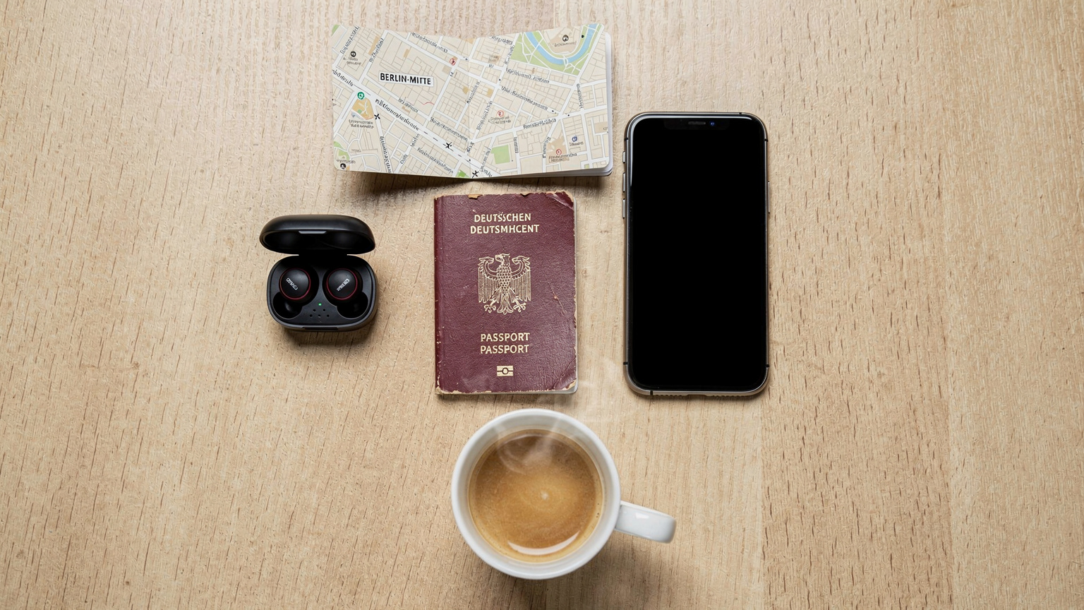 Eine Flat-Lay-Ansicht von Reisepass, Smartphone, Kopfhörern, einer kleinen Karte von Berlin-Mitte und einer dampfenden Tasse Kaffee auf einem Holztisch, alles bereit für eine Reiseplanung.