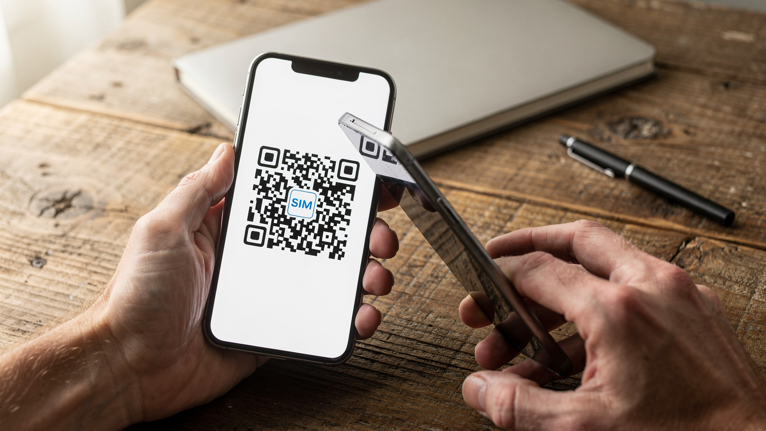 Nahaufnahme von Händen, die ein Smartphone halten, während ein QR-Code für eine eSIM-Aktivierung auf einem Holztisch mit einem Notizbuch und einem Kugelschreiber gescannt wird.