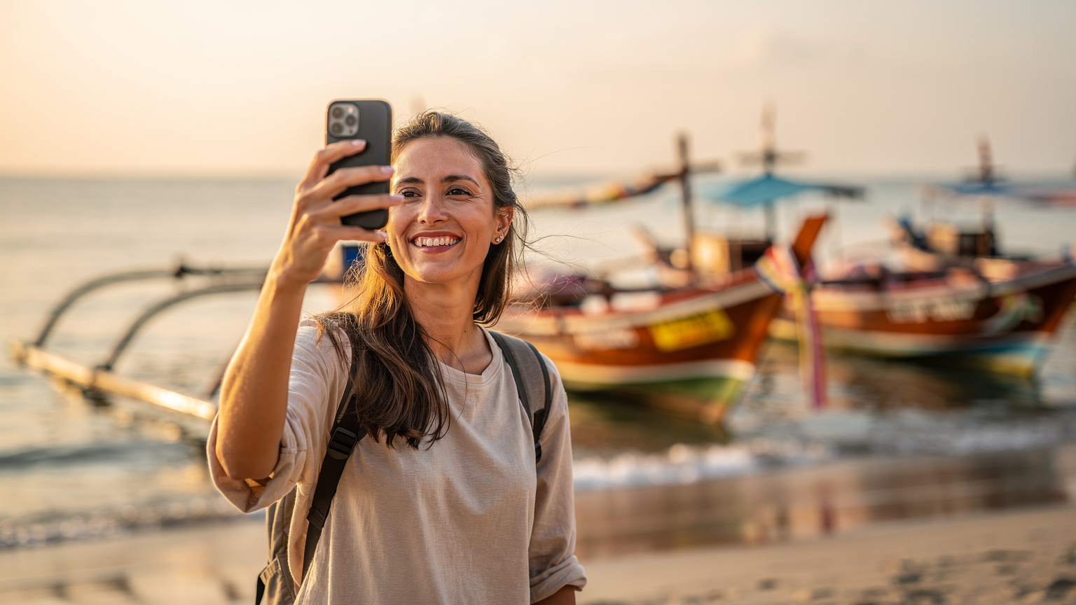 Seorang pengembara sedang tersenyum sambil melakukan panggilan video di kawasan pantai di Sanur, Bali, Indonesia, menikmati sambungan internet yang lancar. Di latar belakang, kelihatan perahu-perahu nelayan yang berwarna-warni.