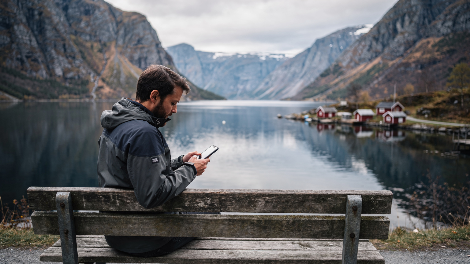 Ein Reisender sitzt auf einer Holzbank mit Blick auf einen norwegischen Fjord, während er sein Smartphone hält und sich die atemberaubende Landschaft ansieht.