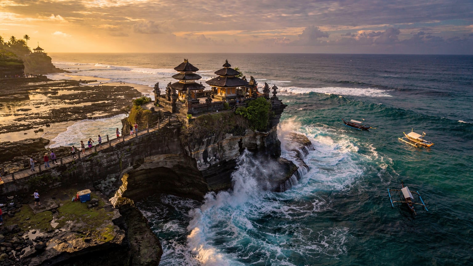 巴厘岛海神庙(Tanah Lot Temple)的标志性航拍全景, 金色的夕阳余晖洒在寺庙和波涛汹涌的海面上, 几艘小船点缀其间, 远处有游客的身影。