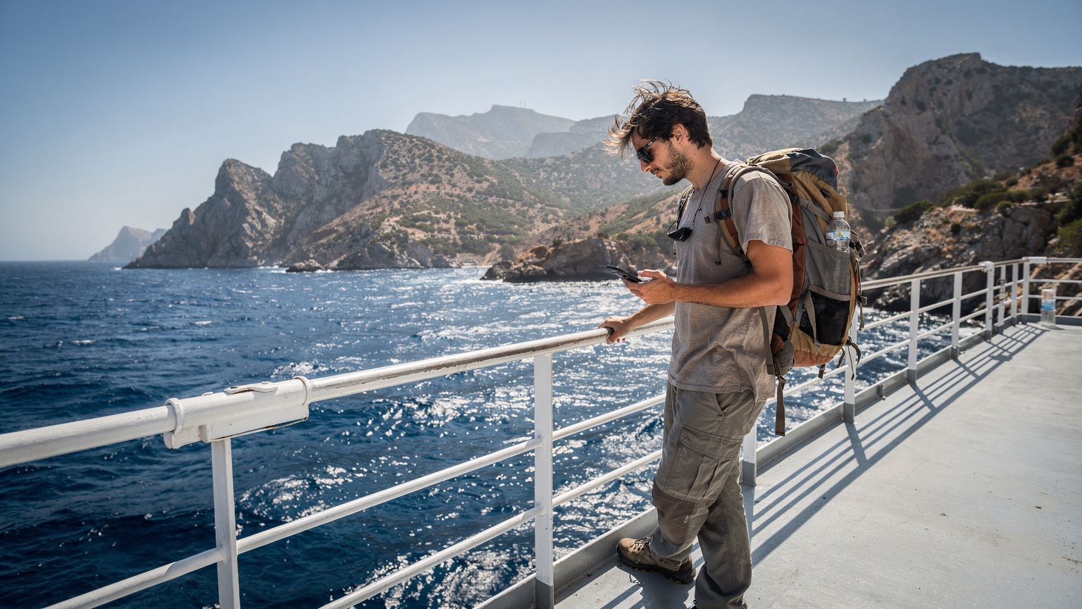 Un jeune homme au look de baroudeur consulte son téléphone à bord d'un ferry moderne traversant la mer Méditerranée, avec le littoral accidenté de la Corse ou de la Sardaigne en arrière-plan.