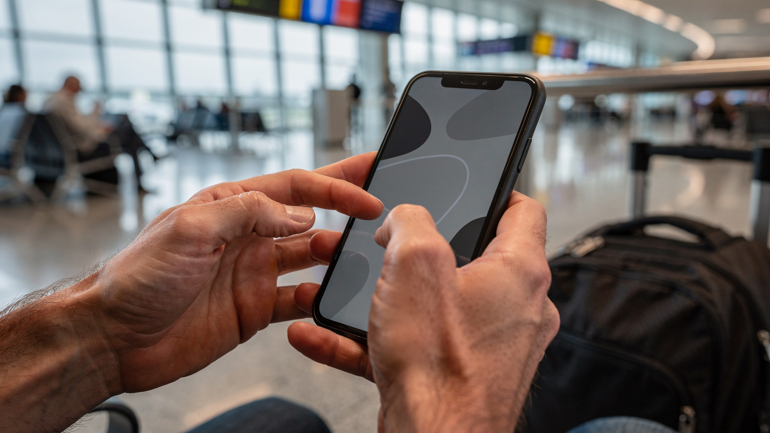Gros plan sur des mains qui téléchargent une carte hors ligne sur un smartphone, avec un sac à dos de voyage en arrière-plan, dans un hall d'aéroport animé.