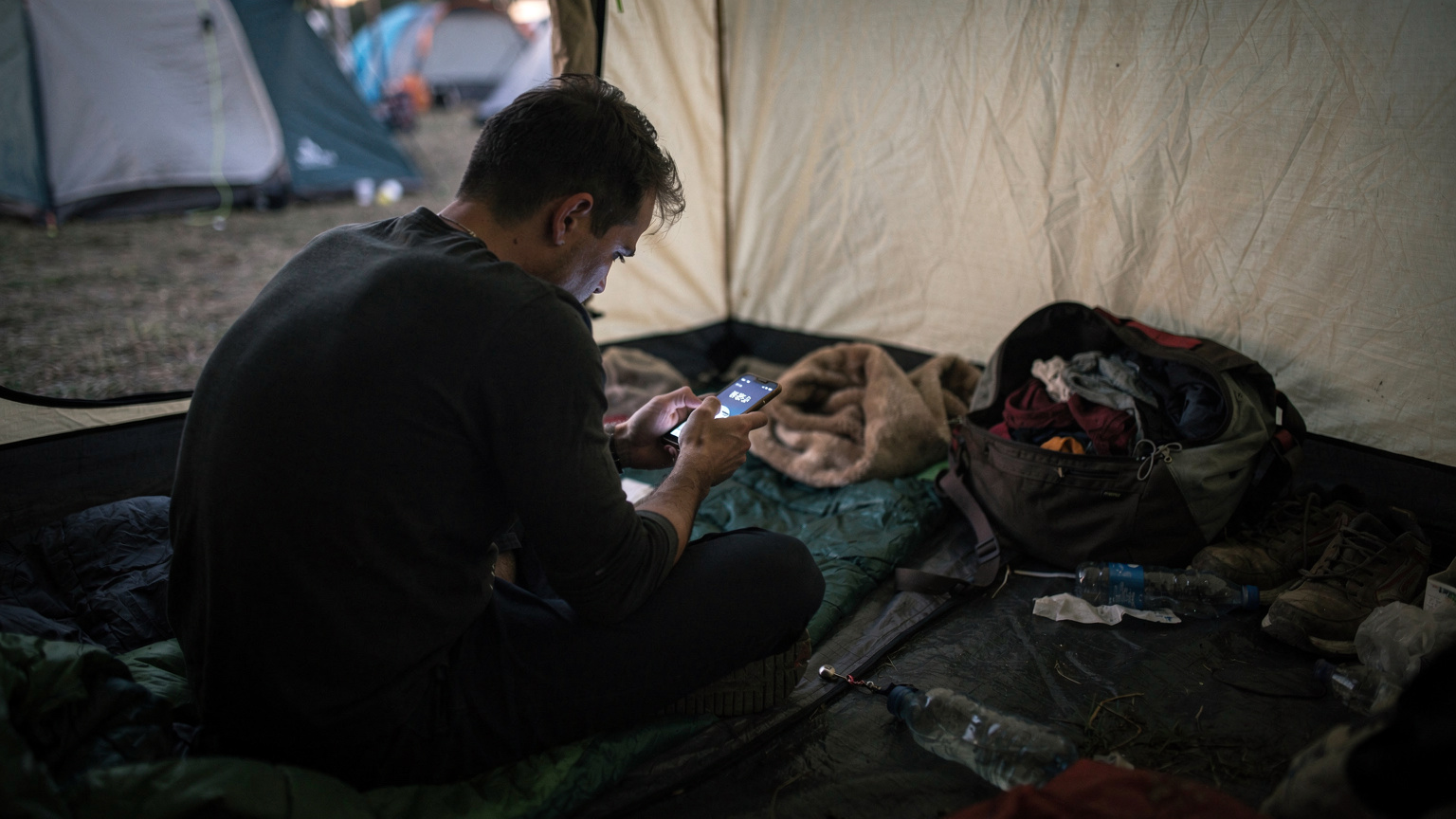 En person, ansiktet bortvänt, sitter i ett trångt tält på en festivalcamping och försöker desperat få mobiltäckning på sin smartphone, med en svag signalindikator på skärmen.