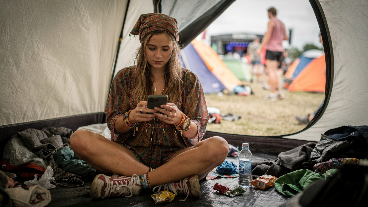 En tjej i färgglada festival kläder tittar på sin smartphone i en tältstad på Roskilde festival, omgiven av andra tält och människor i bakgrunden.