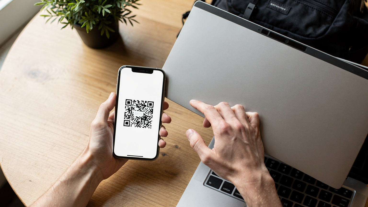 Close-up overhead shot of a traveler's hands holding a smartphone displaying a Cellesim QR code, while a laptop's webcam focuses on the code, initiating an eSIM activation process in a bright hostel common area.