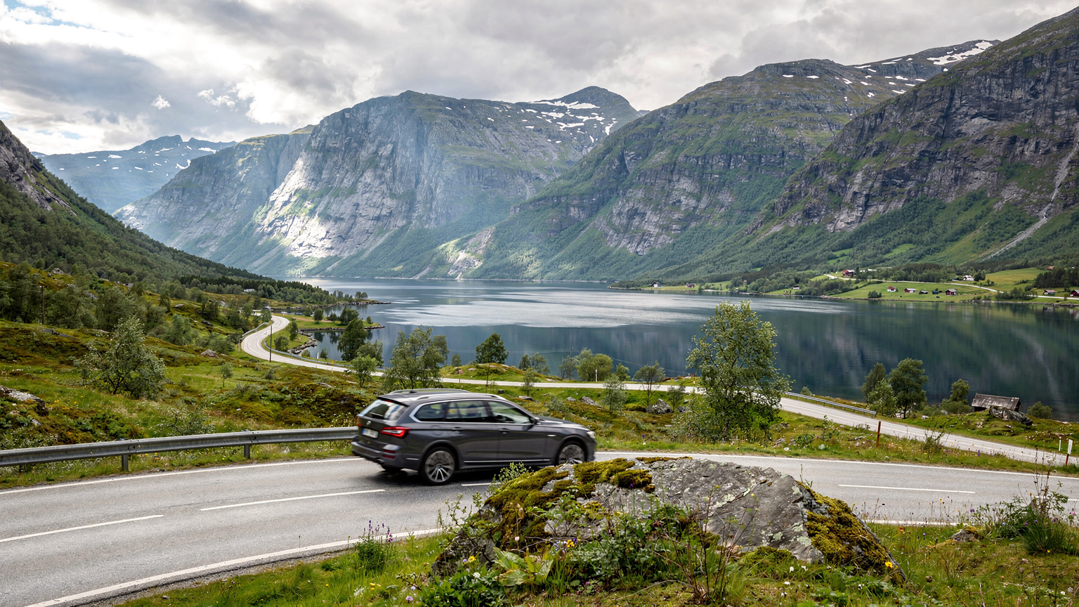 Een gezinsauto rijdt over een schilderachtige weg in Noorwegen, omringd door weelderige groene bergen en een glinsterend fjord onder een helderblauwe hemel.