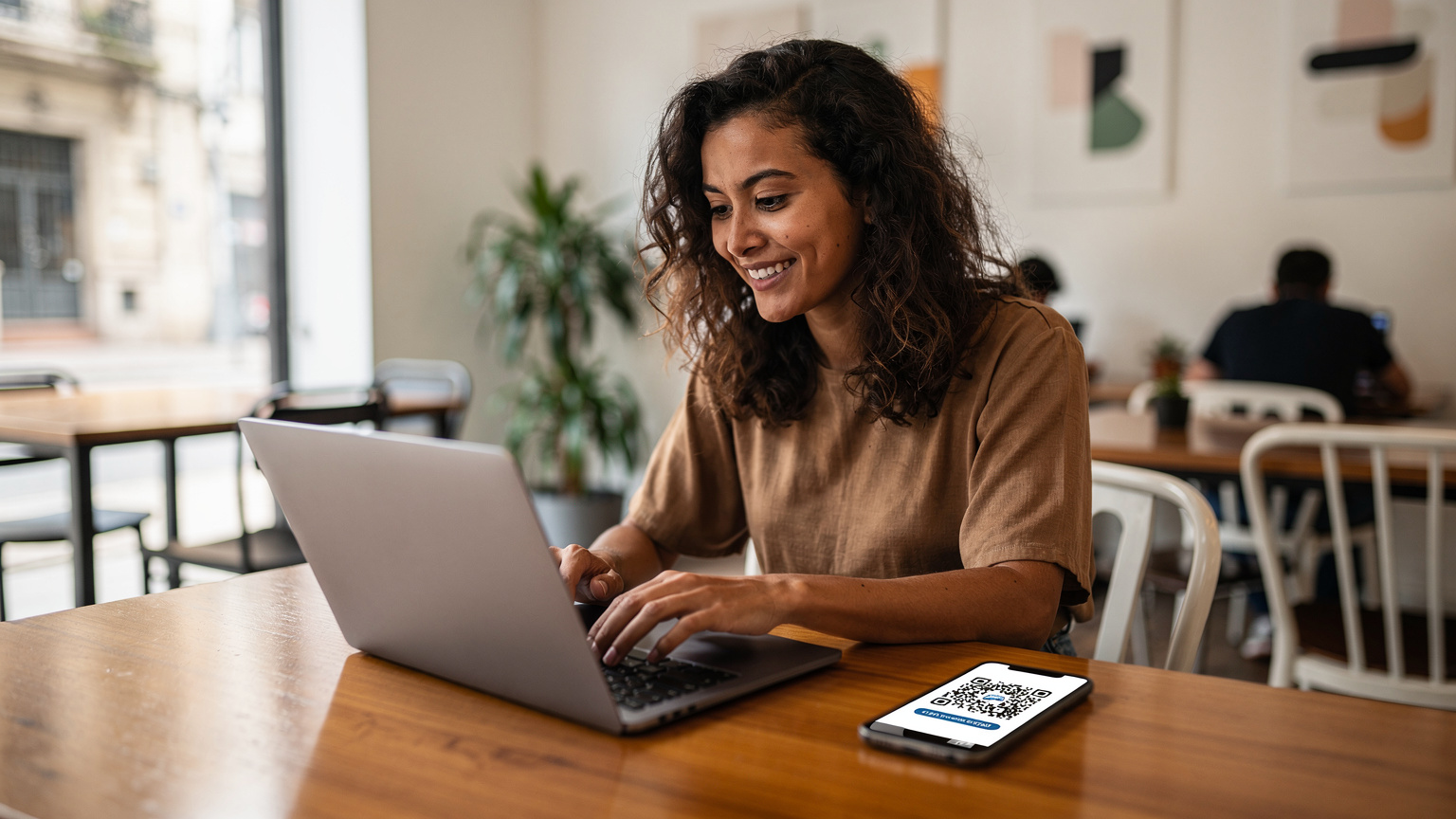 Uma mulher sorridente com cabelos cacheados, sentada em uma mesa de madeira em um co-working moderno no bairro de Palermo, Buenos Aires, Argentina, digitando em seu laptop com um celular mostrando um QR code de eSIM ao lado. Uma xícara de café está na mesa.